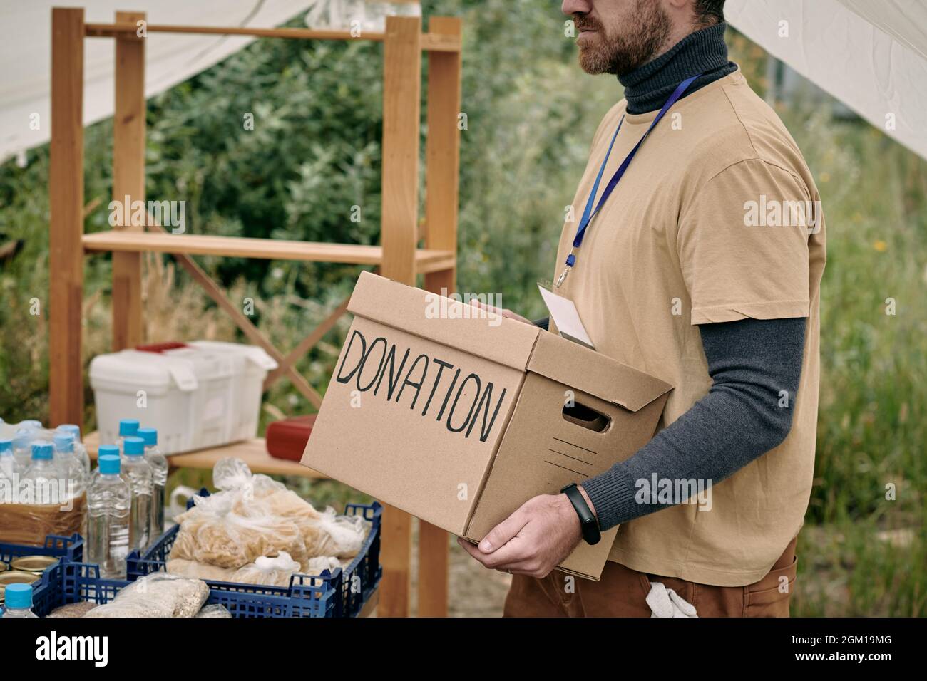 Volontaire barbu avec badge portant une boîte en carton avec don à la table avec des marchandises pour sans-abri Banque D'Images