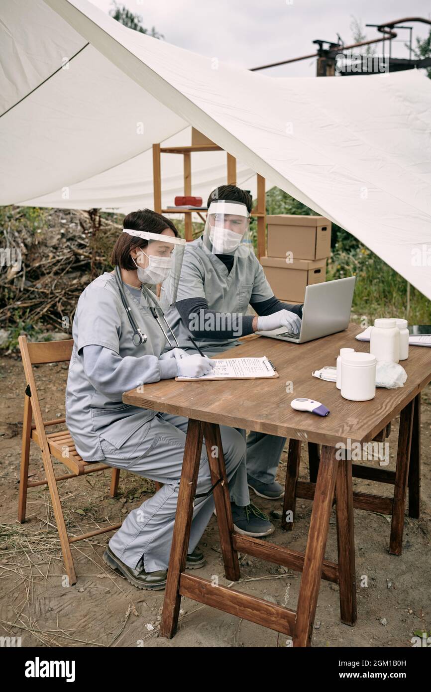 Équipe de médecins en sécurité masque assis à une table dans une tente et travaillant avec des cartes médicales de réfugiés dans une zone éloignée Banque D'Images