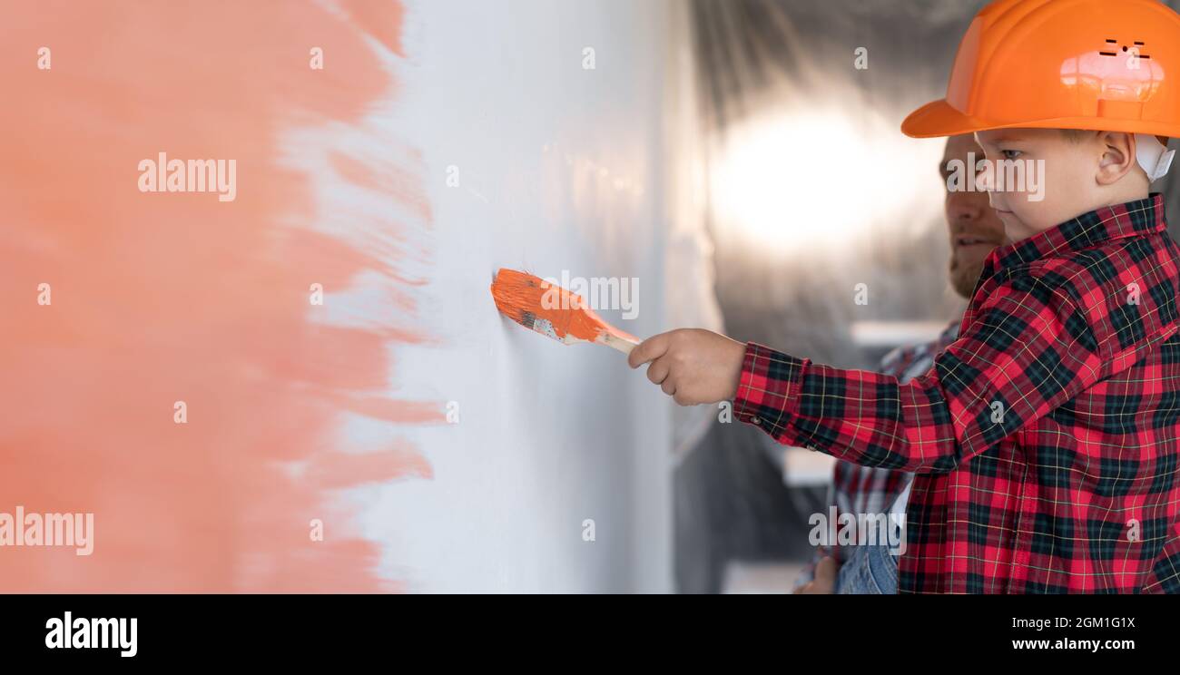 Vue de profil d'un garçon dans un casque avec papa peint le mur dans la chambre en orange. La famille fait des réparations à domicile. Travail pour hommes et mur coloré. Banque D'Images
