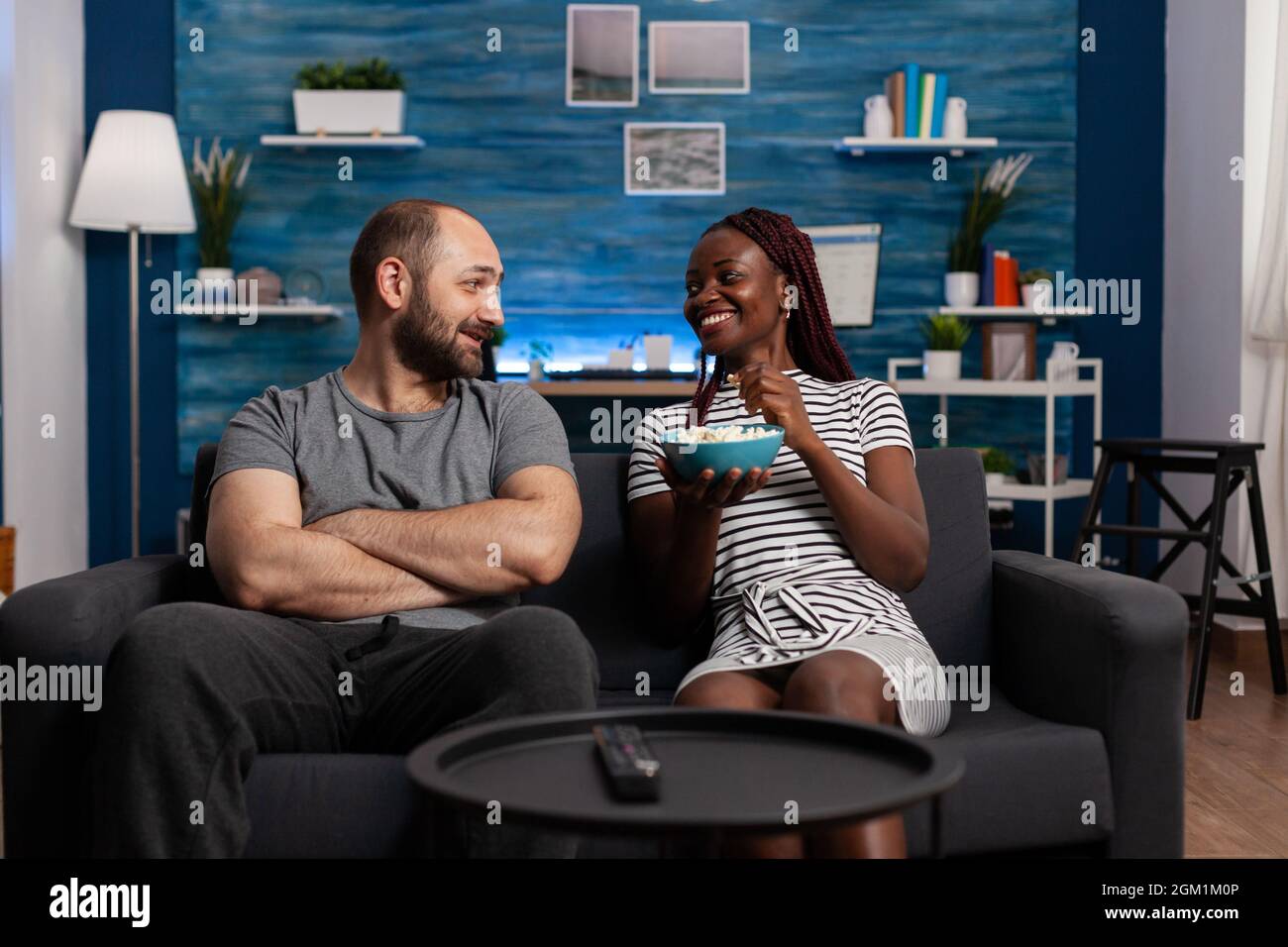 Un jeune couple interracial rit et bavarde sur son canapé. POV de partenaires multiethniques souriant tout en ayant pop-corn et de discuter de film à la télévision. Des gens de race mixte qui profitent de temps libre Banque D'Images