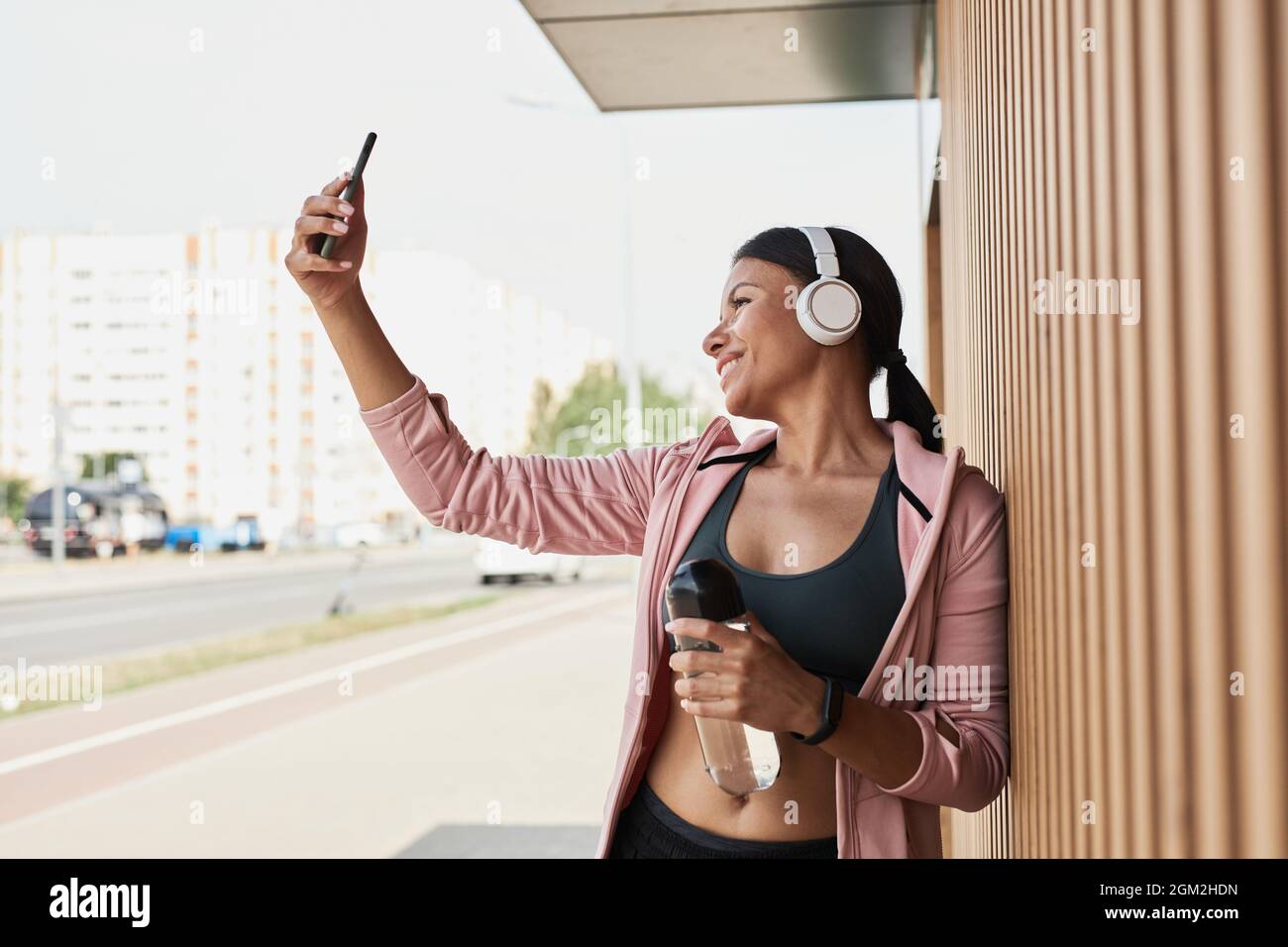 Jeune sportif en casque sans fil souriant à l'appareil photo tout en faisant un portrait de selfie sur un téléphone mobile à l'extérieur Banque D'Images