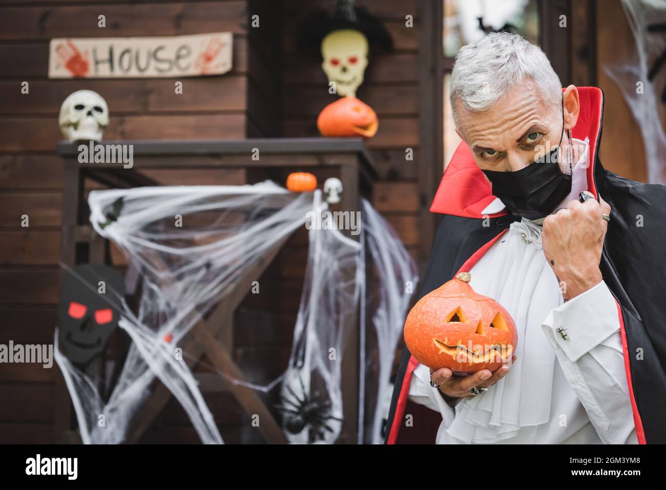 homme d'âge moyen en costume de vampire et masque médical noir près de la maison floue avec décoration d'halloween Banque D'Images