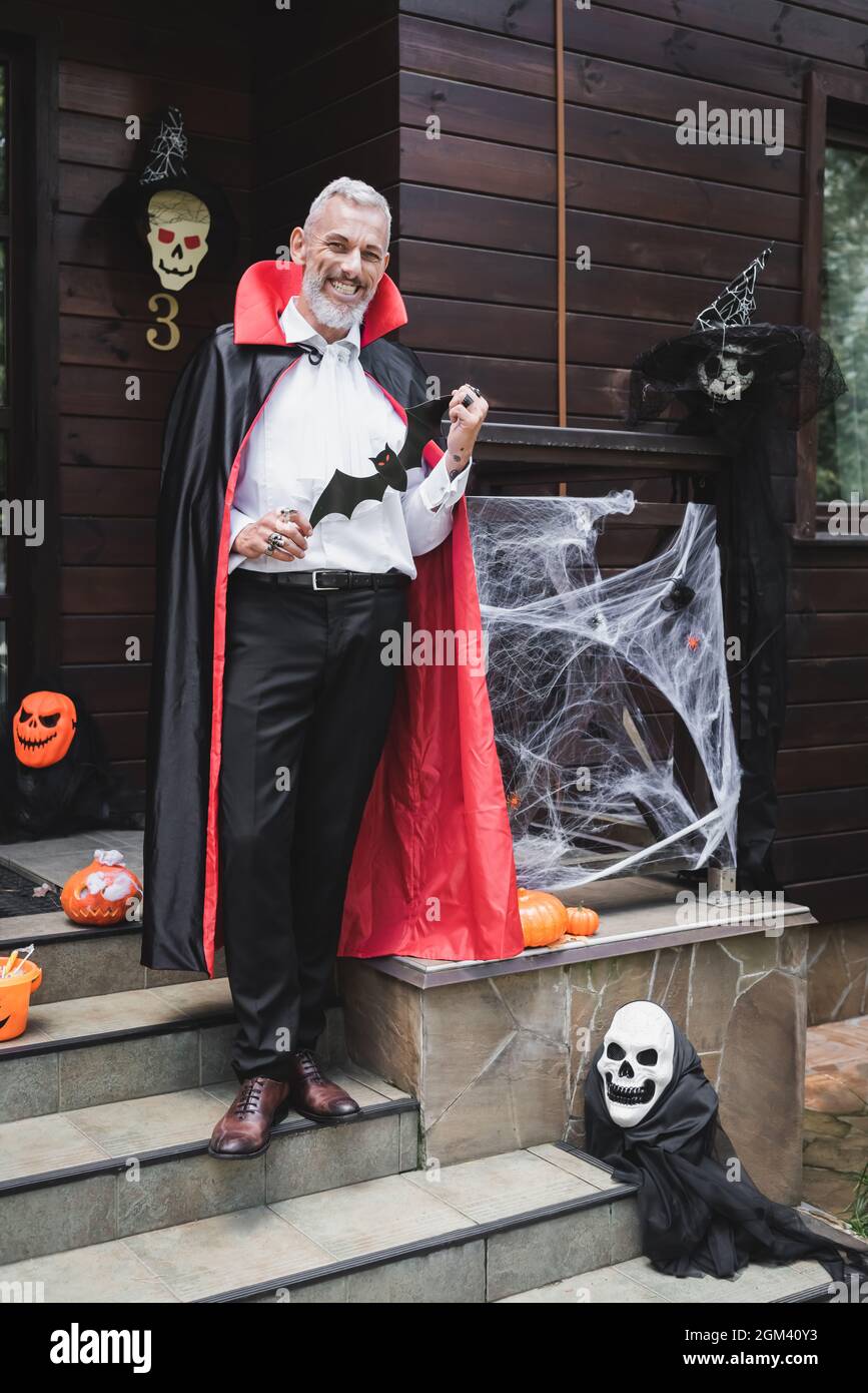 joyeux homme mature en costume de vampire tenant une batte en papier sur le porche avec la décoration d'halloween Banque D'Images