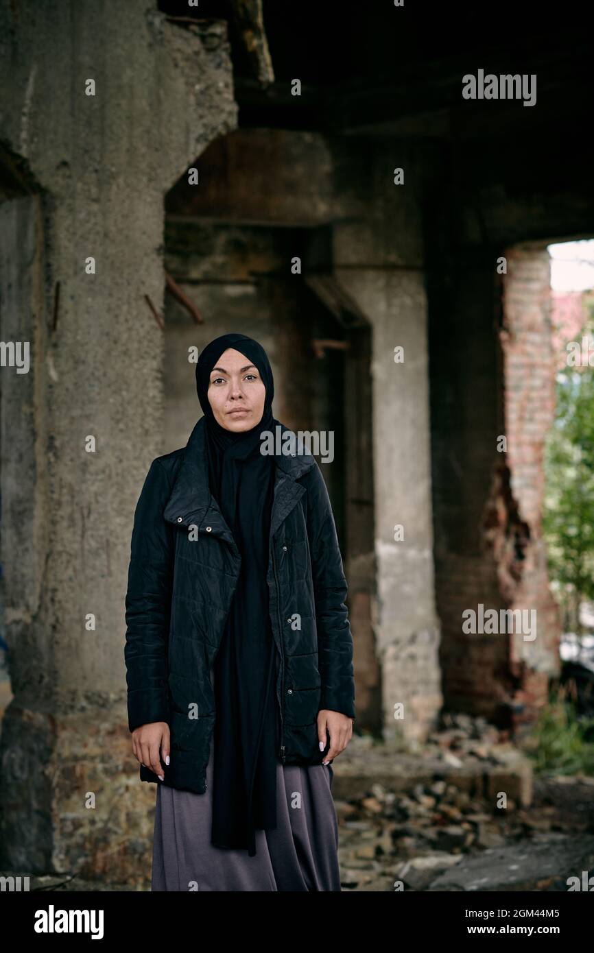 Portrait d'une jeune femme musulmane réfugiée triste dans un hijab et une longue veste debout contre un bâtiment en ruines Banque D'Images