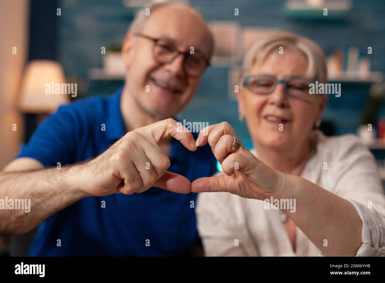 Couple senior créant une figure en forme de coeur avec les mains à la maison. Des personnes âgées mariées dans l'amour faisant signe romantique tout en regardant l'appareil photo dans le salon. Mari et femme montrant un geste d'affection Banque D'Images
