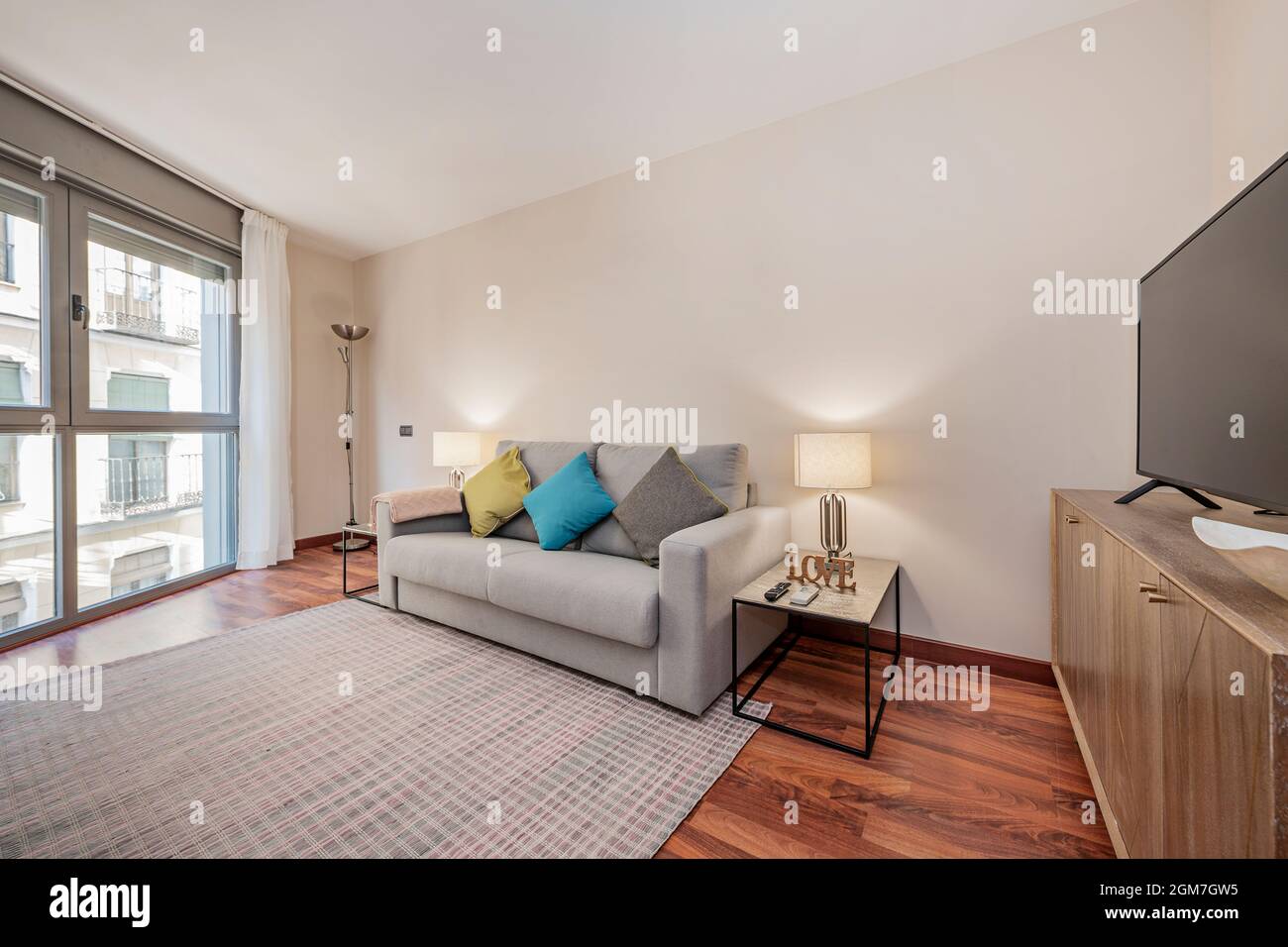 Séjour avec grande fenêtre pleine hauteur, canapé gris avec coussins colorés et grande télévision dans un appartement de location de vacances Banque D'Images