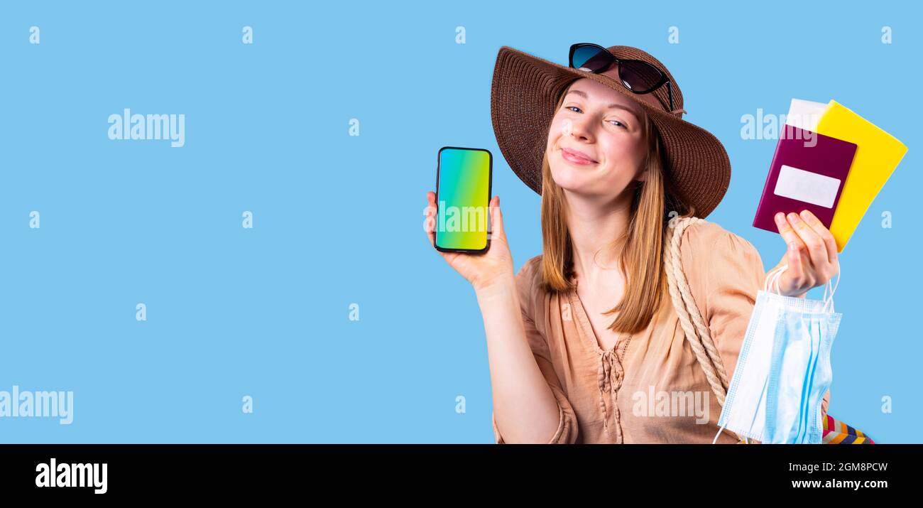 Très heureuse femme touriste portant un chapeau et des lunettes de soleil posant sur l'appareil photo tenant le téléphone mobile, le masque médical et le passeport. Banque D'Images
