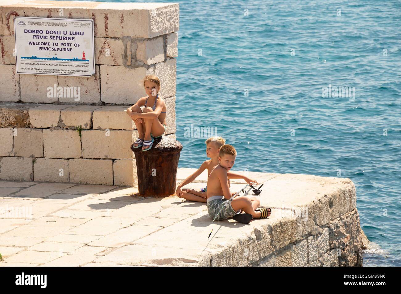 Prvic Sepurin, Croatie - 25 août 2021 : enfants blonds assis au soleil sur la jetée en pierre de pêche et regardant au loin, et une mer derrière Banque D'Images