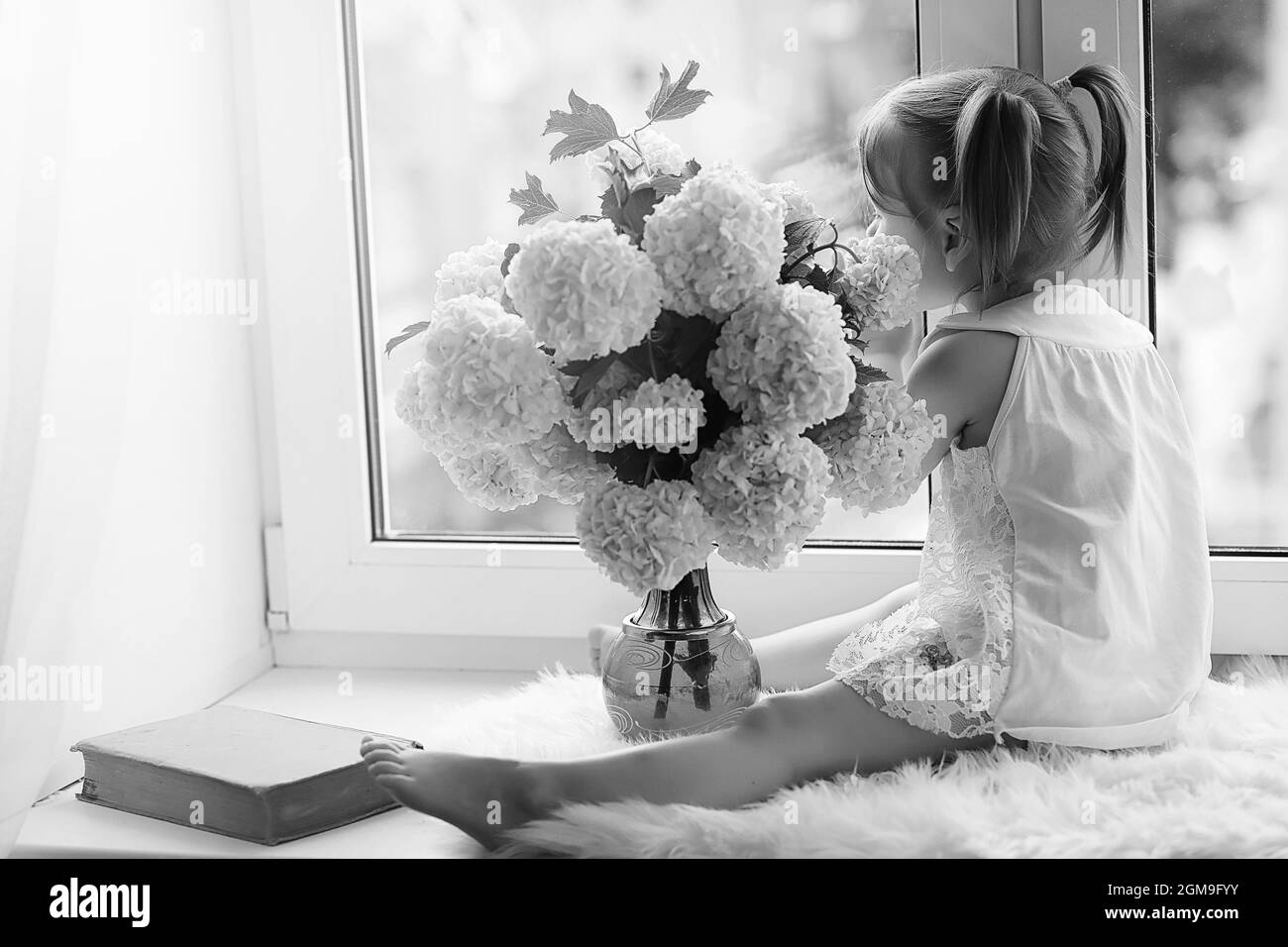 Une petite fille est assise sur le rebord de la fenêtre. Un bouquet de fleurs dans un vase près de la fenêtre et une fille d'essence de fleurs. Une petite princesse dans un dre blanc Banque D'Images