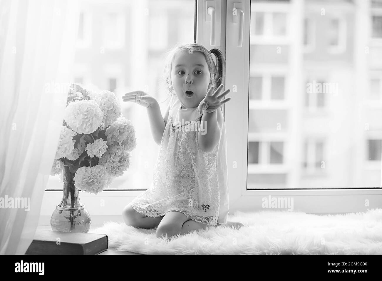 Une petite fille est assise sur le rebord de la fenêtre. Un bouquet de fleurs dans un vase près de la fenêtre et une fille d'essence de fleurs. Une petite princesse dans un dre blanc Banque D'Images