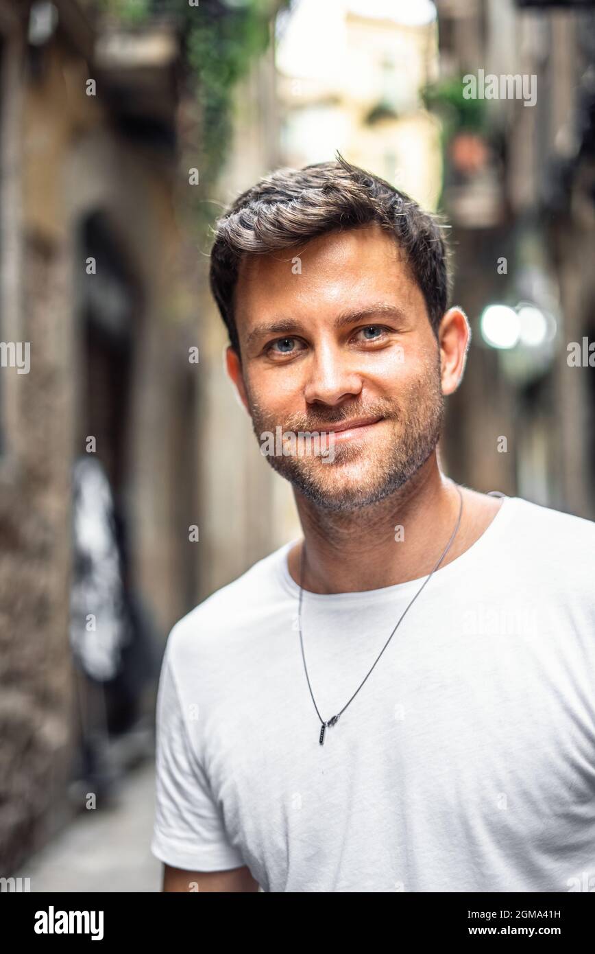 Contenu jeune homme barbu dans un t-shirt blanc décontracté souriant et regardant l'appareil-photo tout en se tenant sur la petite rue de la vieille ville en été Banque D'Images