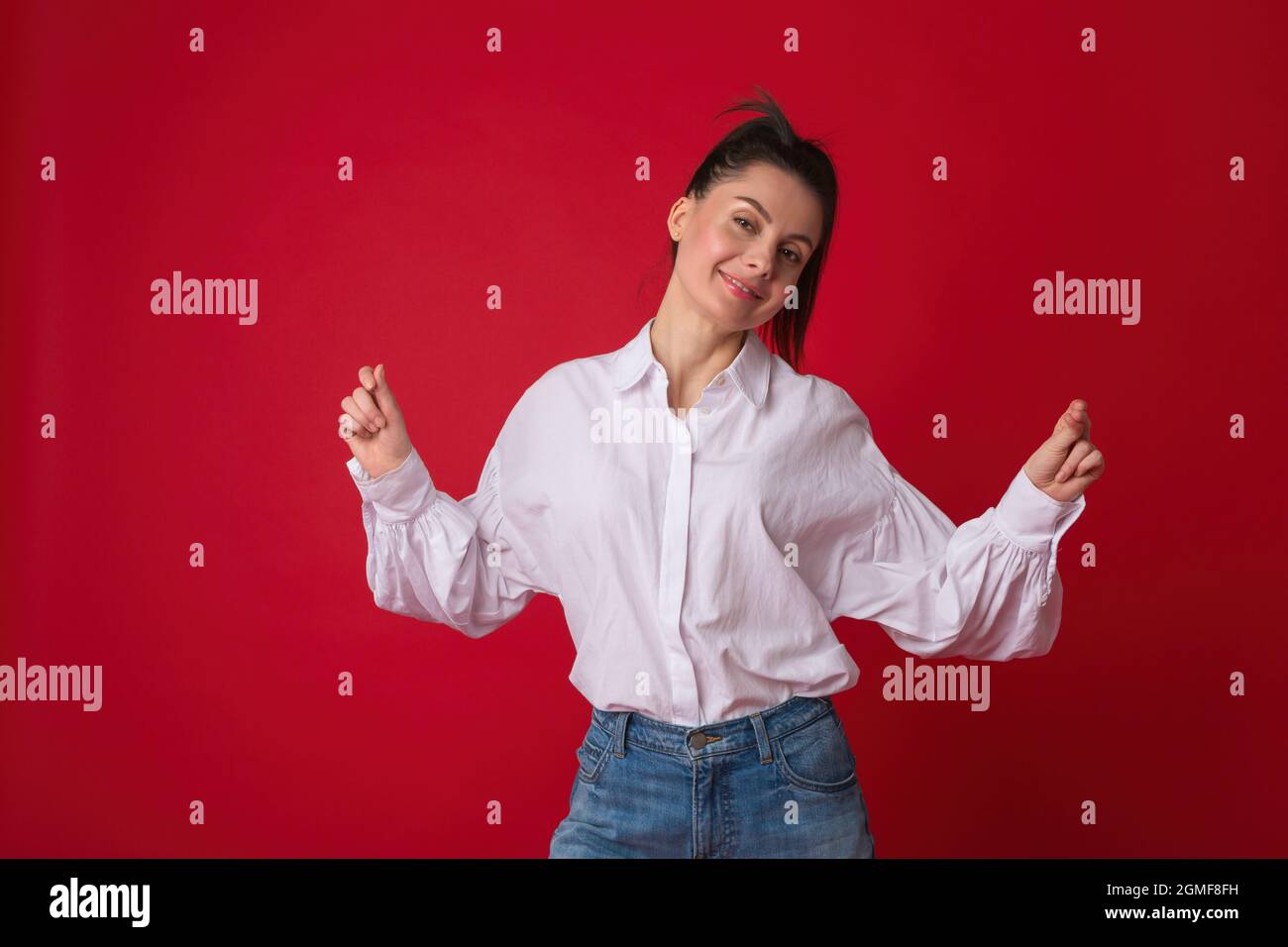 Bonne jeune femme dansant avec le sourire. Belle femme portant une chemise blanche décontractée et un Jean bleu classique posé en studio sur fond rouge Banque D'Images