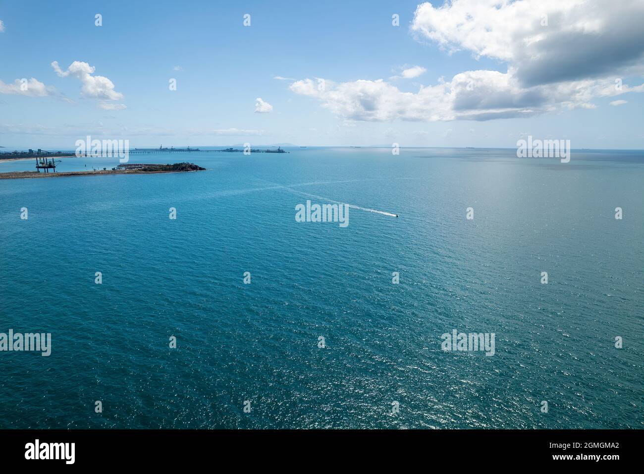 Un petit bateau à moteur se dirige à travers l'eau d'un port d'exportation de charbon vers la rive sous un ciel bleu ciel nuageux avec le soleil qui gécoute sur l'eau et un di Banque D'Images