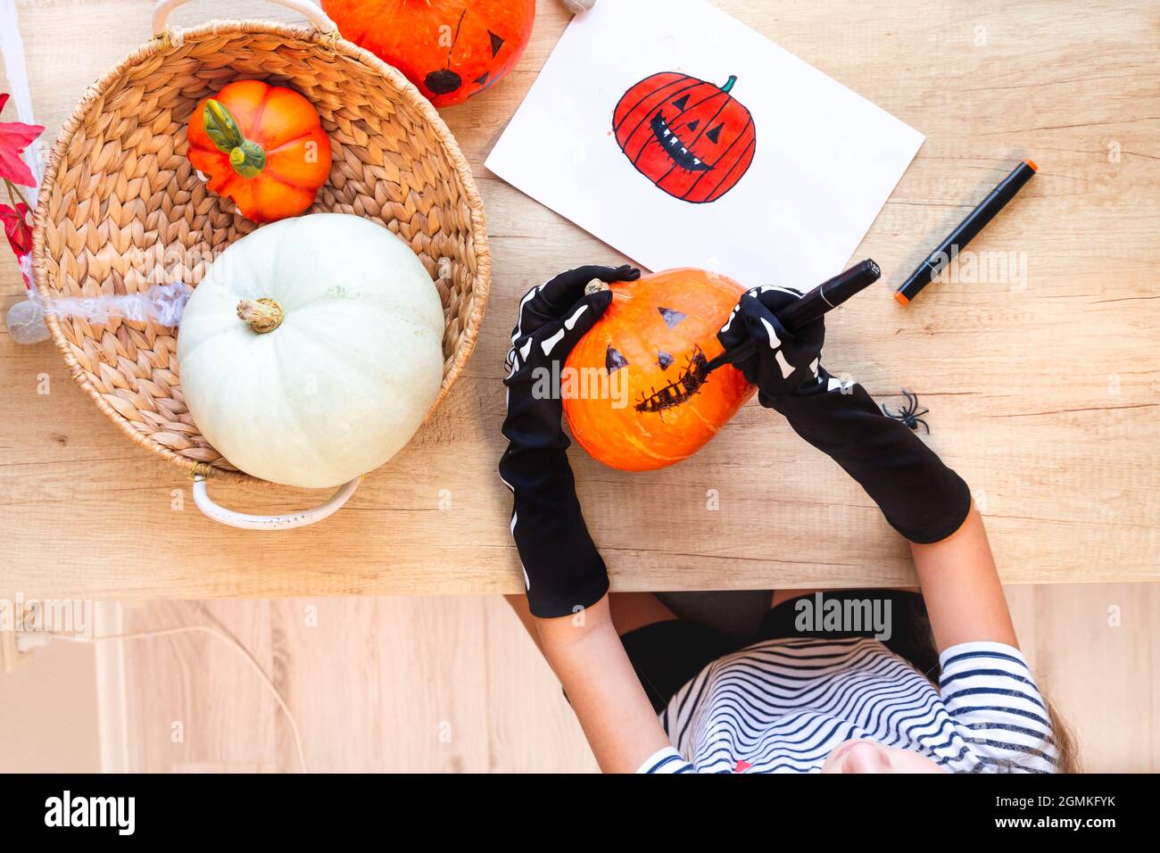 Préparation pour les vacances, la décoration de la maison pour Halloween. Un enfant en gants noirs tire un visage riant sur une citrouille, assis à un bureau, à côté d'un Banque D'Images