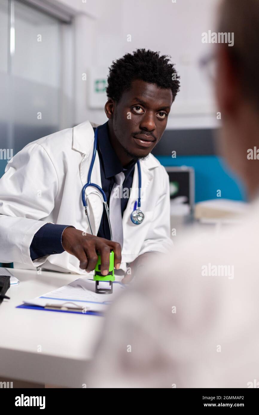 Medic of african american ethnicity Holding Stamp pour signer des documents pour le patient assis à son bureau. Homme noir avec profession de médecin utilisant l'outil sur les dossiers papier pour le chekup à la clinique de santé Banque D'Images