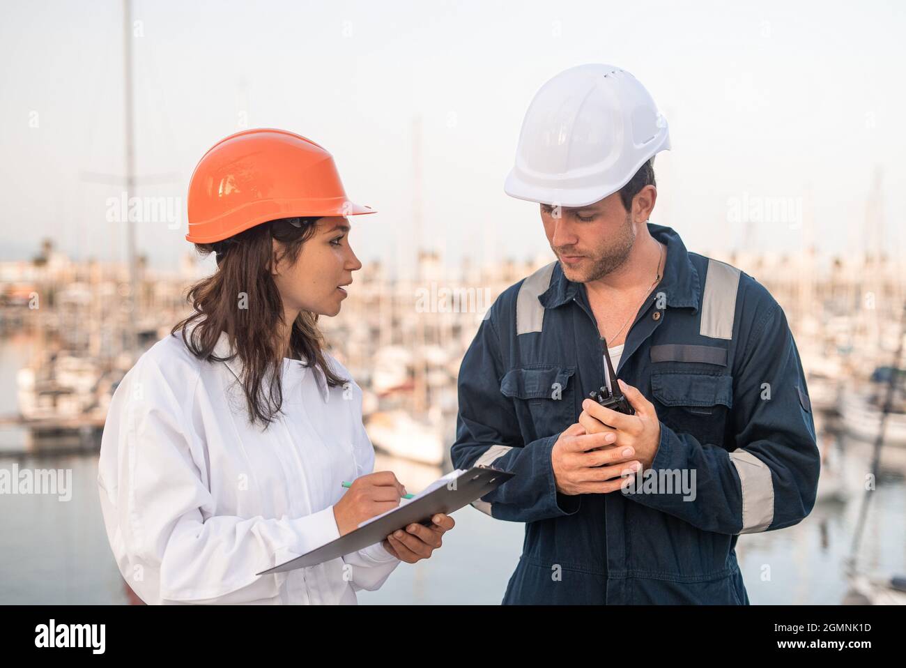 Inspecteur femme professionnel remplissant la liste de contrôle sur le presse-papiers tout en communiquant avec l'employé mâle avec les mains de la radio VHV pendant l'inspection du port de mer Banque D'Images