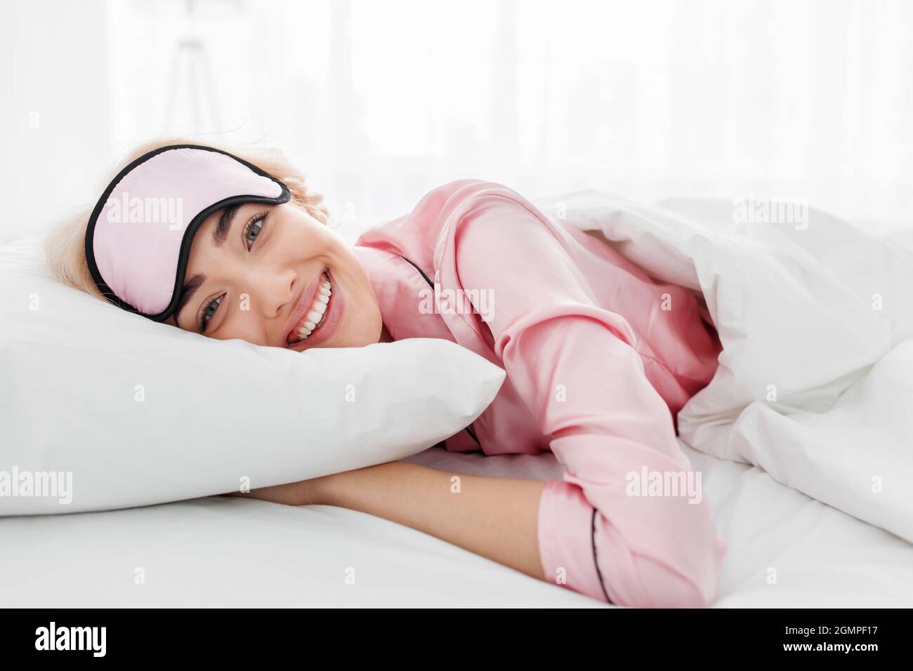 Souriant jolie jeune femme blonde caucasienne en pyjama rose et masque de sommeil appréciant le matin Banque D'Images