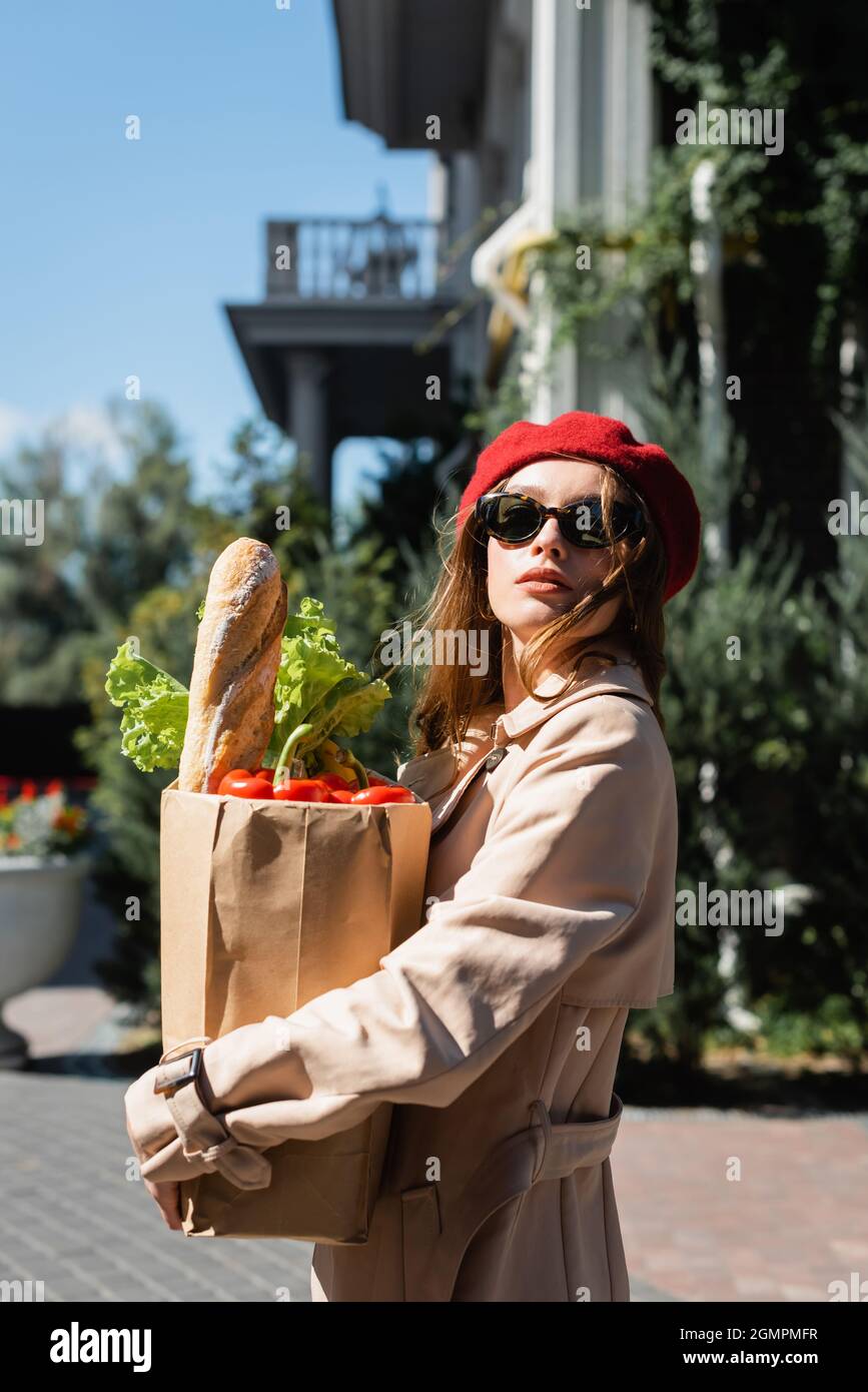 jeune femme en trench-coat beige, lunettes de soleil et béret rouge tenant un sac en papier avec des articles d'épicerie Banque D'Images