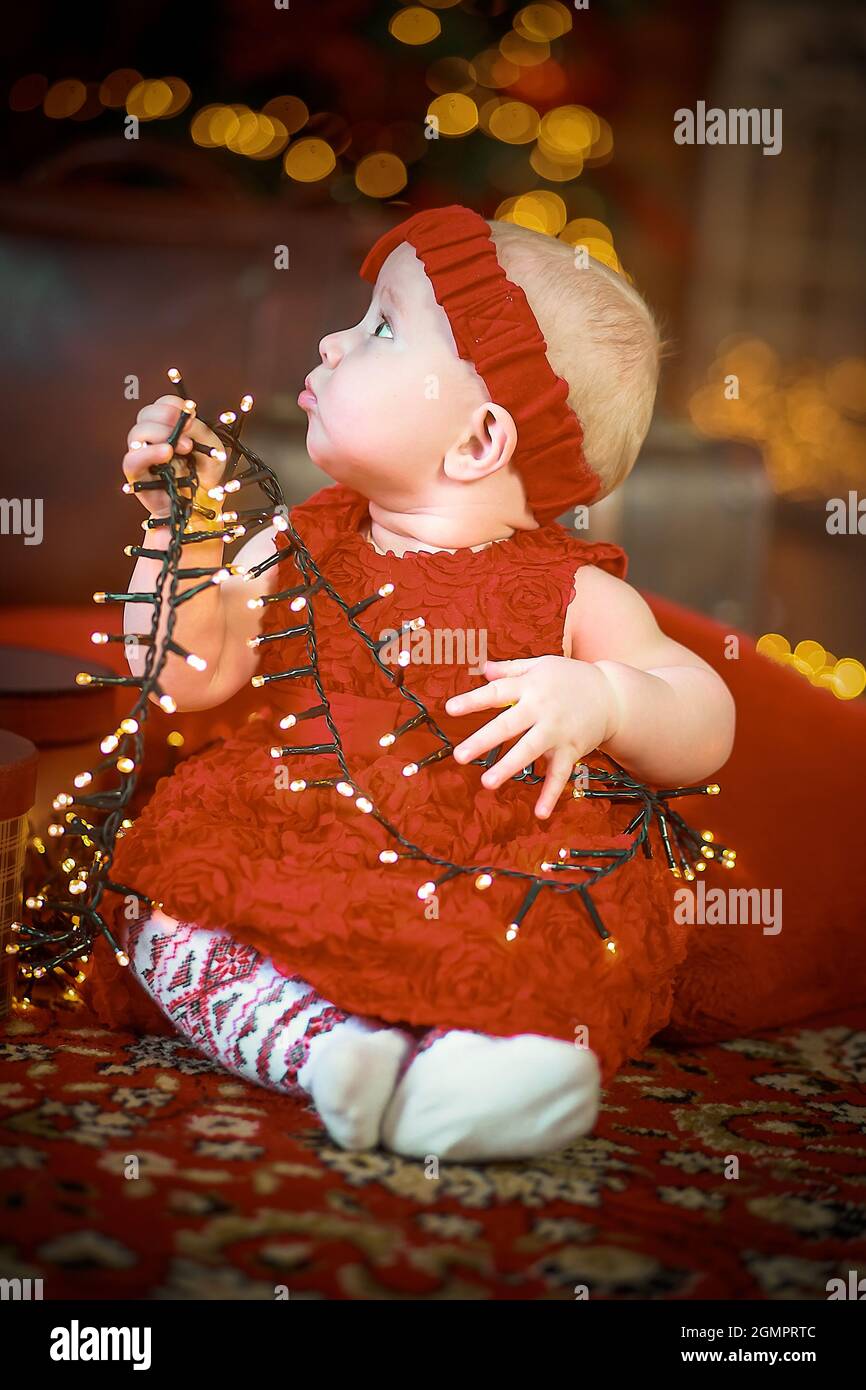 Petite fille en robe rouge à l'arrière-plan de Noël arbre de Noël est titulaire d'garland en mains. bébé 6 mois fête Noël. Banque D'Images