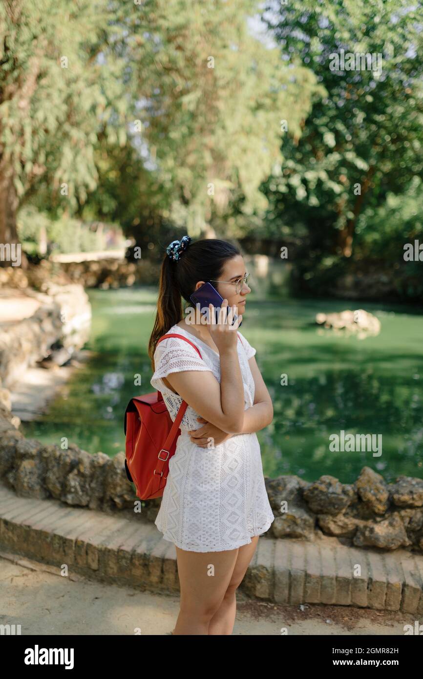 Photo moyenne d'une jeune femme avec une robe blanche et un sac à dos rouge parlant au téléphone dans un parc Banque D'Images