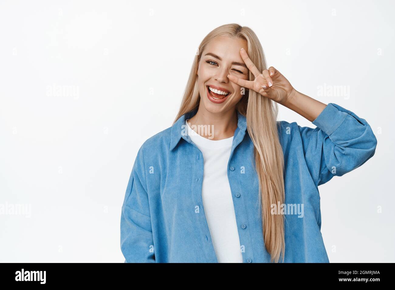 Positive blonde fille montre la paix, le V-signe geste contre les yeux et wencing happiy, souriant avec des dents blanches à la caméra, debout contre l'arrière-plan de studio Banque D'Images