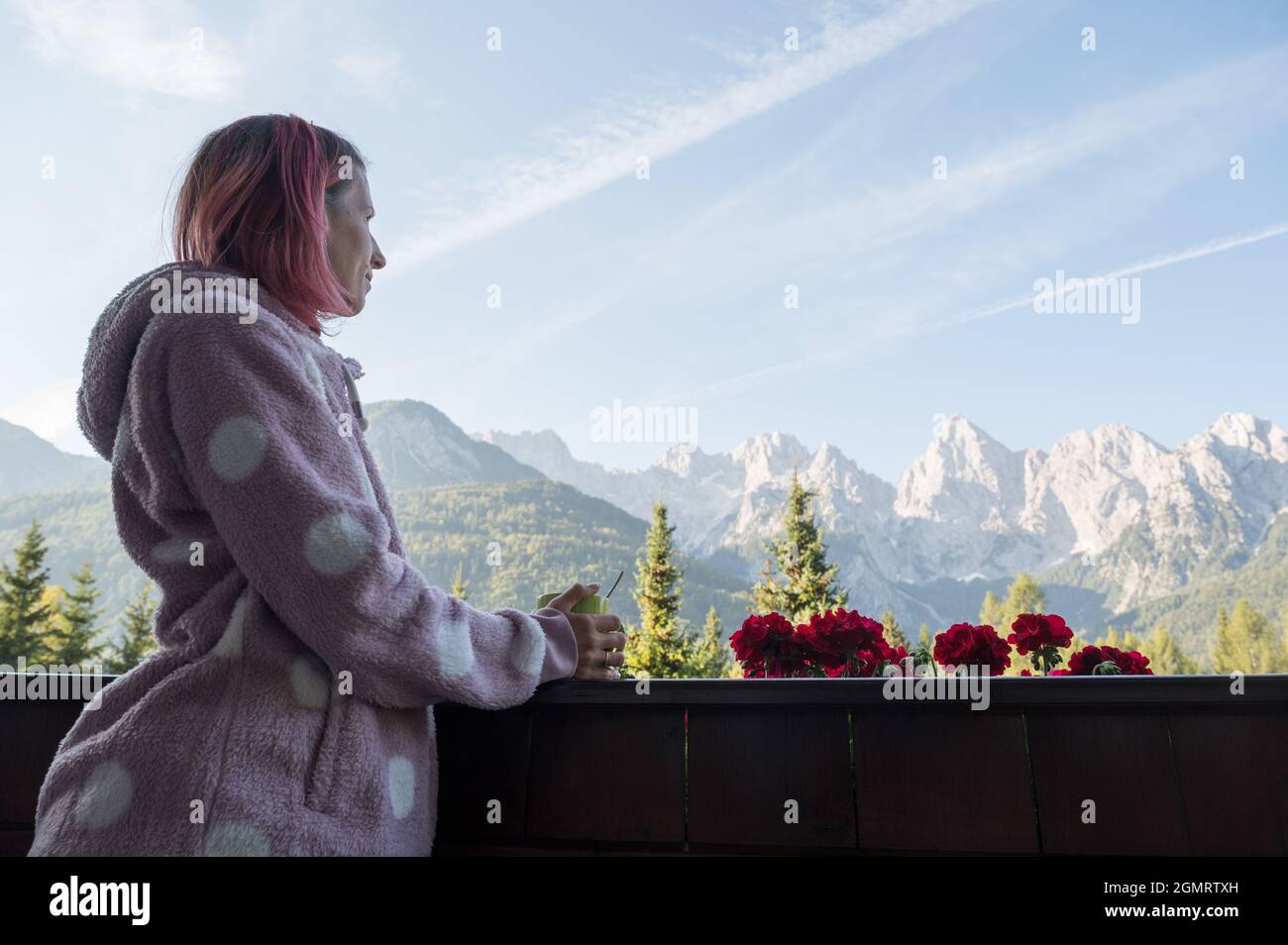Jeune femme aux cheveux roses dans un confortable chandail chaud appuyé sur une clôture de balcon en bois, tenant une tasse de café et profitant de la vue magnifique sur les montagnes. Banque D'Images