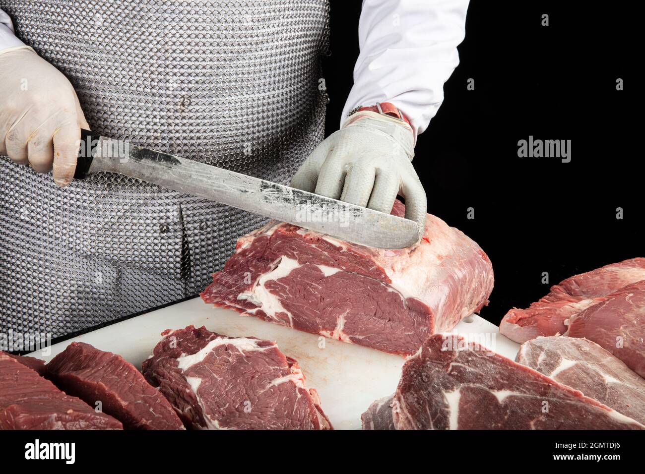 Gros plan des morceaux de viande hachés et des mains de boucher dans des gants spéciaux coupant avec un couteau. Viande de porc ou de bœuf sur la table du boucher. Le travailleur en tout Banque D'Images