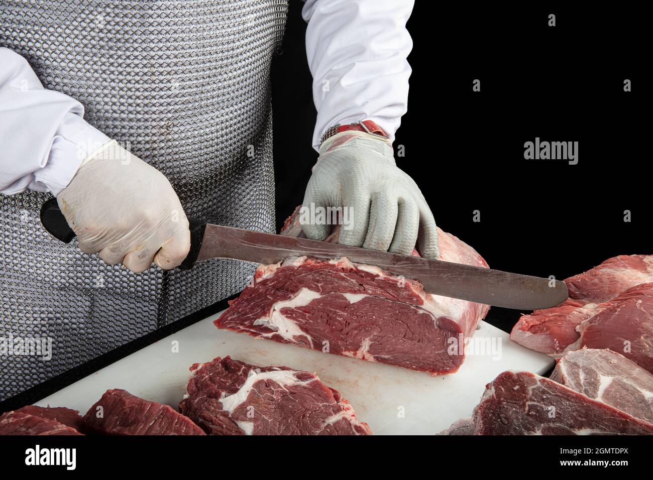 Gros plan des morceaux de viande hachés et des mains de boucher dans des gants spéciaux coupant avec un couteau. Viande de porc ou de bœuf sur la table du boucher. Le travailleur en tout Banque D'Images