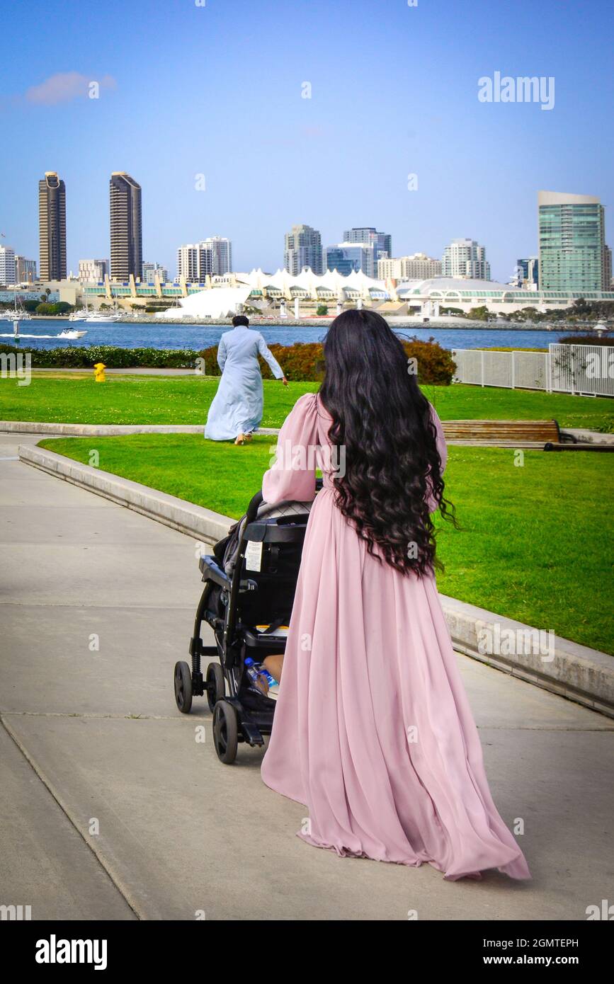 Vue arrière des femmes avec de longs cheveux noirs portant une longue robe rose poussant une poussette le long du chemin de Coronado Ferry Landing à San Diego via la baie, CA Banque D'Images