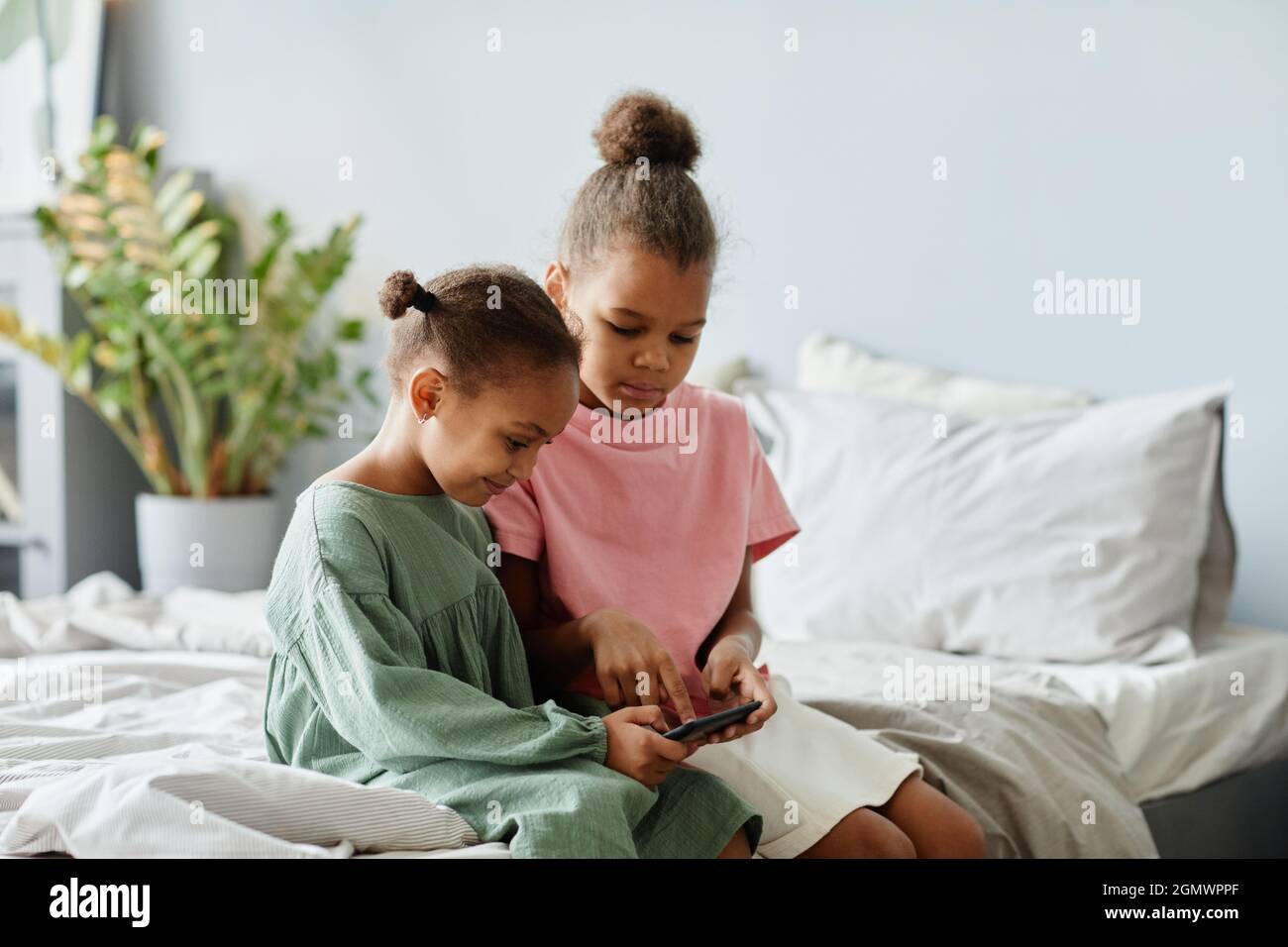 Portrait de deux jolies filles afro-américaines utilisant un smartphone ensemble tout en étant assis sur un lit dans une pièce confortable, espace de copie Banque D'Images