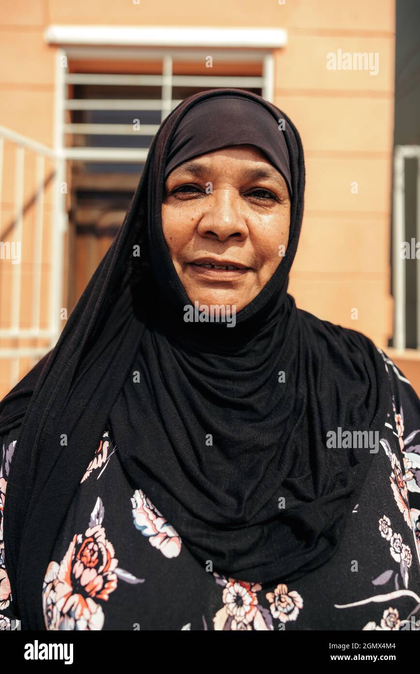 Femme musulmane insouciante regardant la caméra tout en se tenant devant sa maison pendant la journée. Une femme d'âge moyen confiante porte un hijab et un s. Banque D'Images Femme musulmane insouciante regardant la caméra tout en se tenant devant sa maison pendant la journée. Une femme d'âge moyen confiante porte un hijab et un s. Banque D'Images