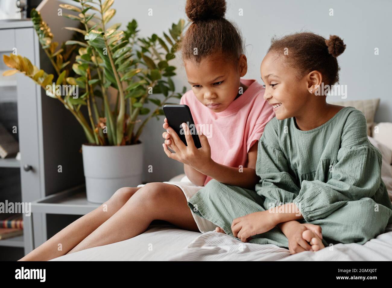 Portrait de deux sœurs mignonnes utilisant un smartphone ensemble tout en étant assis sur un lit dans une pièce confortable, espace de copie Banque D'Images