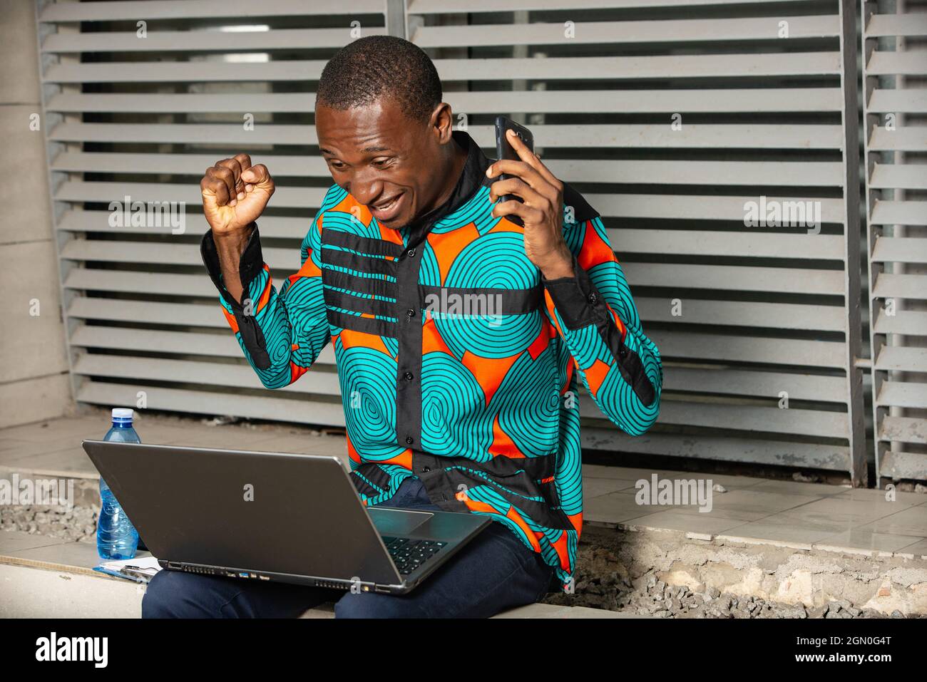 beau jeune africain assis et travaillant avec un ordinateur portable, heureux et excité, faisant des gestes avec les bras levés, souriant et criant pour le succès Banque D'Images