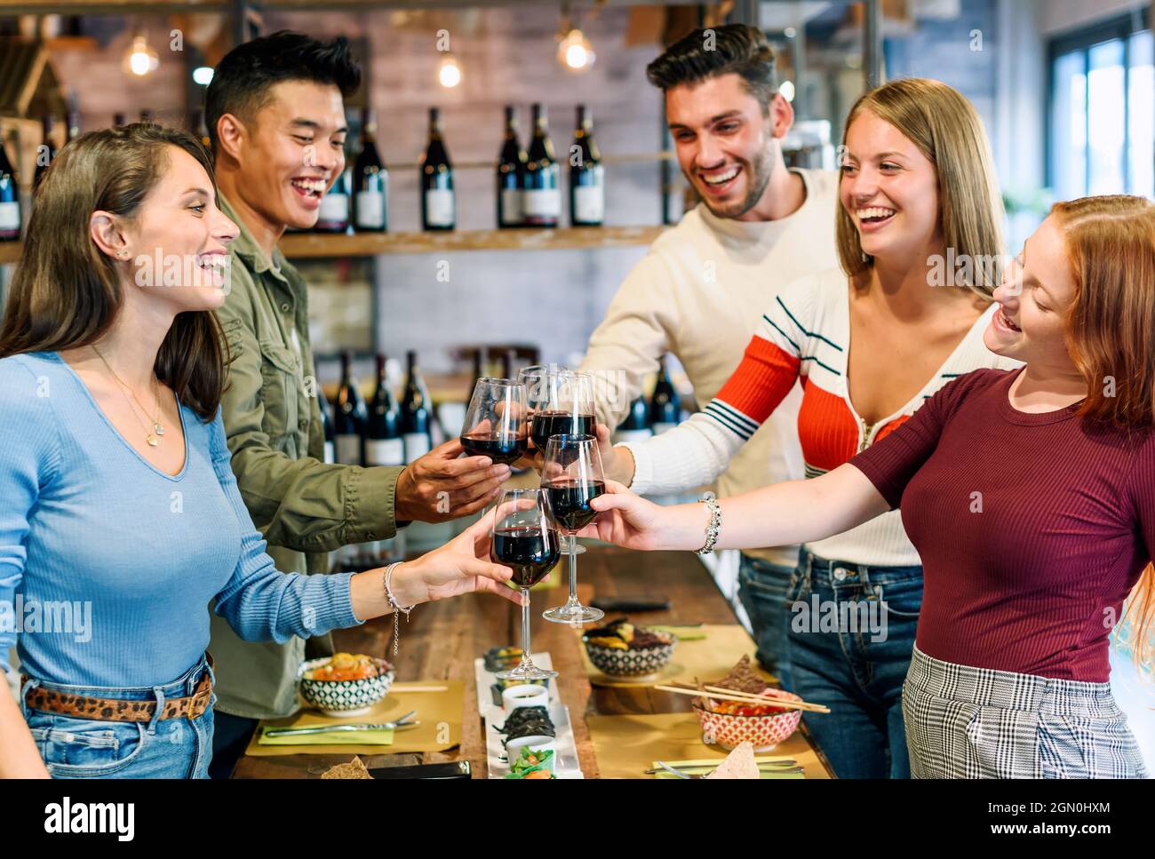 Un groupe de jeunes amis heureux et enthousiastes partageant un toast ensemble en se cligeant dans leurs verres de vin rouge tout en dégustant un repas dans un pub ou un restaurant Banque D'Images