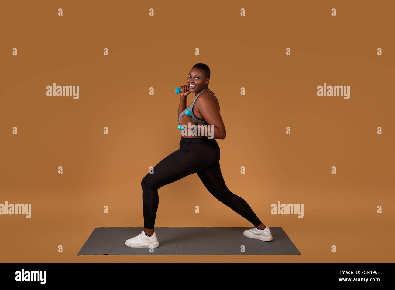 Concept d'entraînement de force. Bonne femme noire curvy faisant des exercices de fente avec des haltères sur fond brun de studio, debout sur le tapis de yoga, regardant ca Banque D'Images