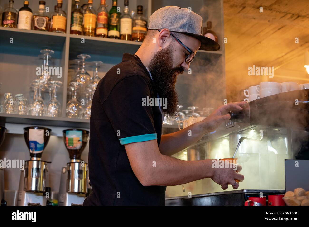 Barista barbu qui prépare de la crème de lait cuite à la vapeur dans un pot en acier, à l'aide d'une machine à café professionnelle. Préparation d'un café Latte ou d'un cappuccino. Banque D'Images