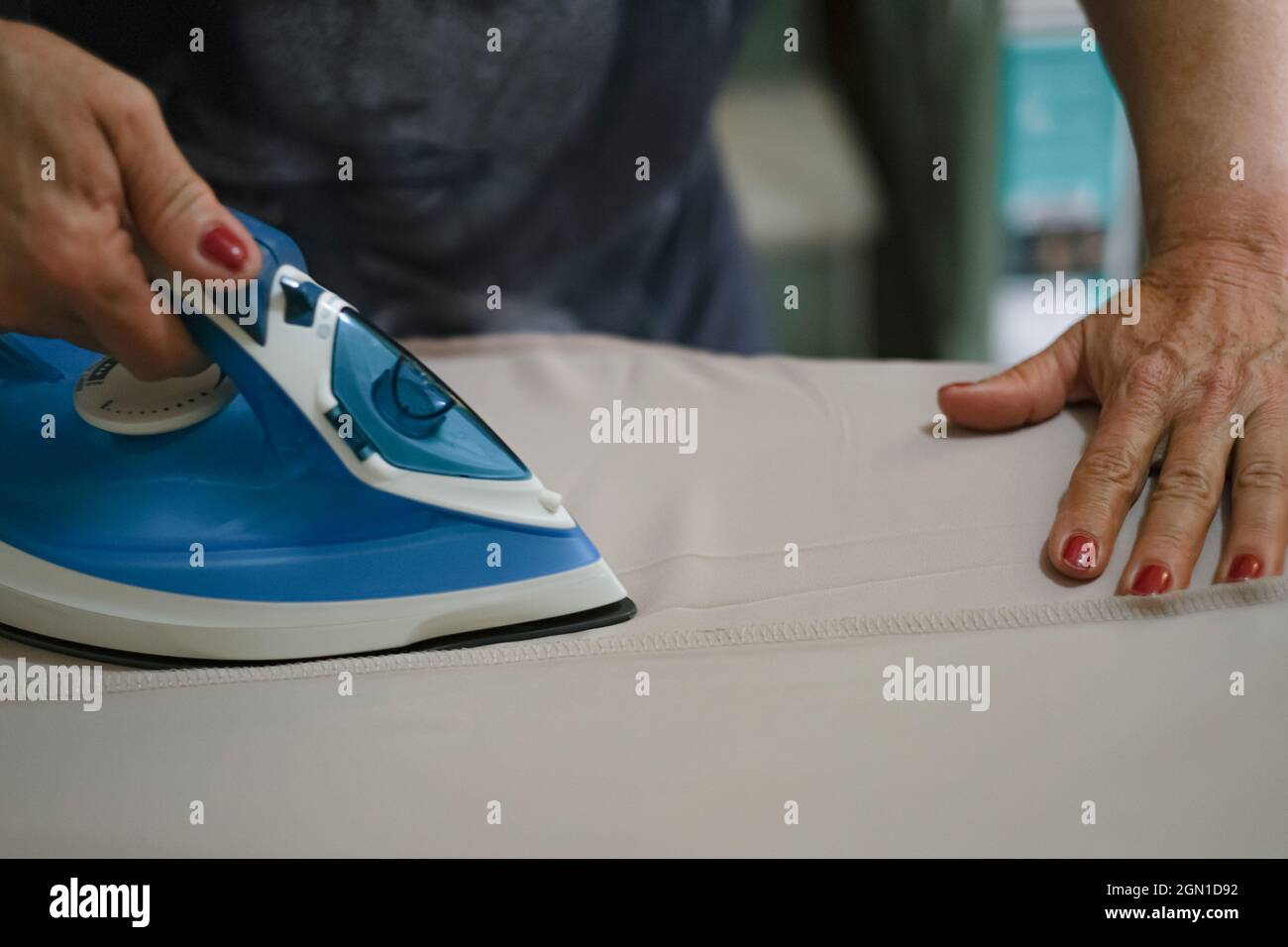 Une femme âgée se tient à la main tout en repassant des vêtements avec un fer à vapeur sur une planche à repasser Banque D'Images