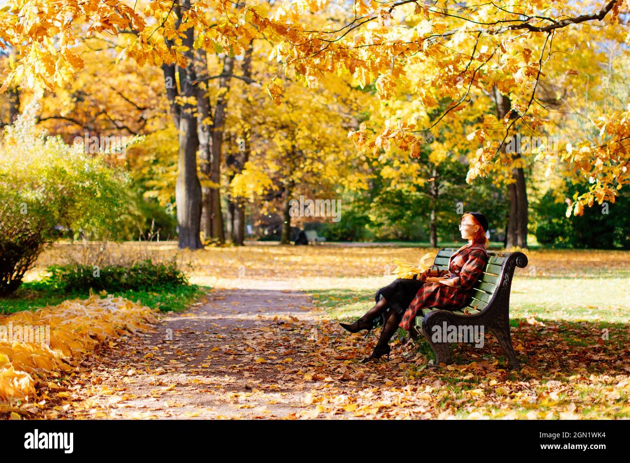 redhead femme en manteau écossais et béret noir assis sur le banc, se reposant dans le parc d'automne à la journée ensoleillée. saison d'automne Banque D'Images