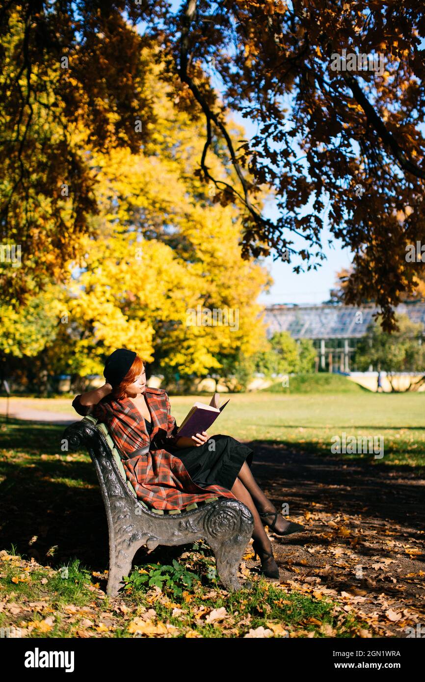 redhead femme en manteau à carreaux et livre de lecture de béret noir sur banc, se reposant dans le parc d'automne à la journée ensoleillée Banque D'Images