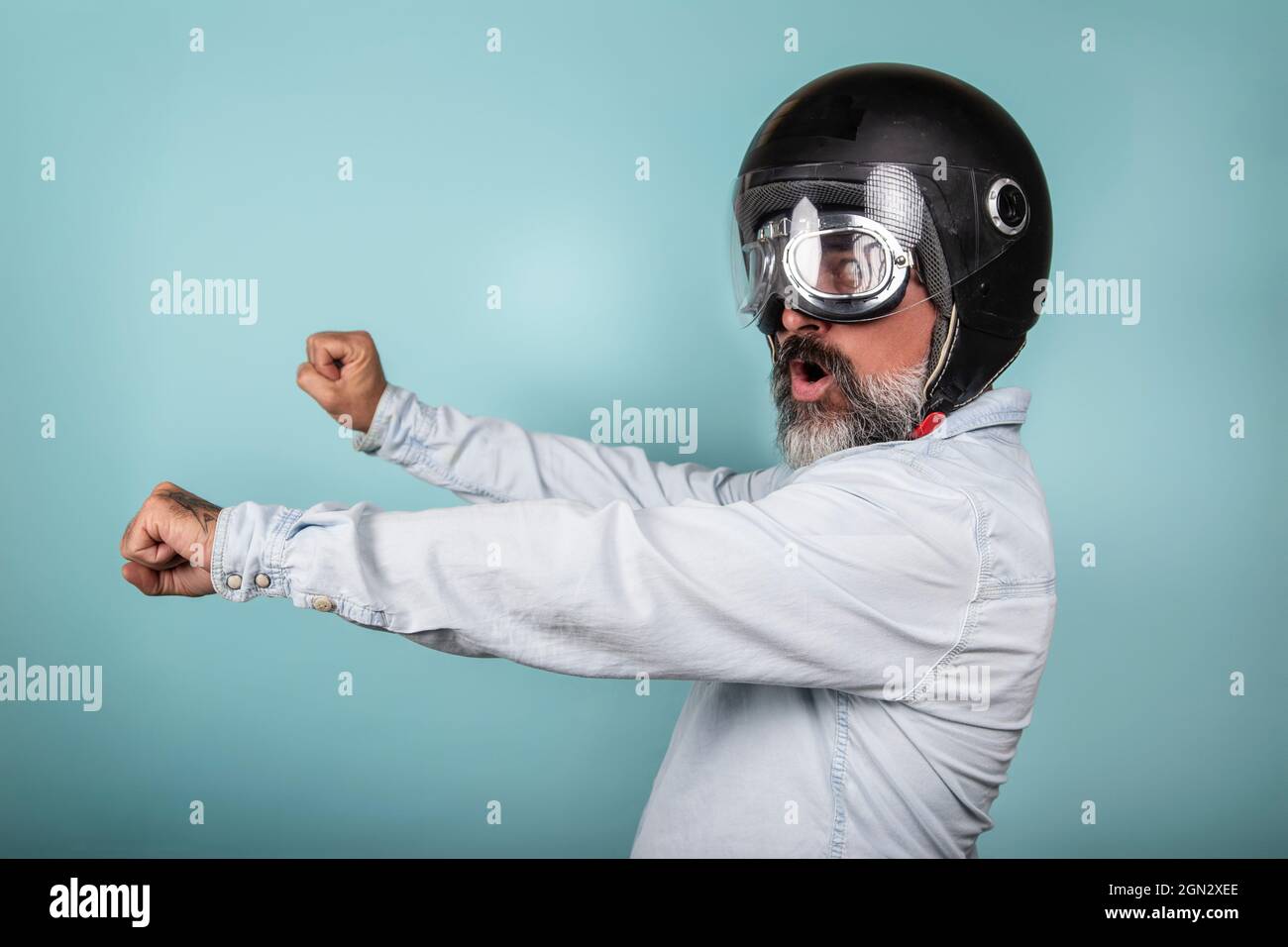 Profil de l'hipster barbu en chemise denim avec casque et lunettes prétendant monter à bord d'une moto, isolé sur un mur bleu de studio. Banque D'Images