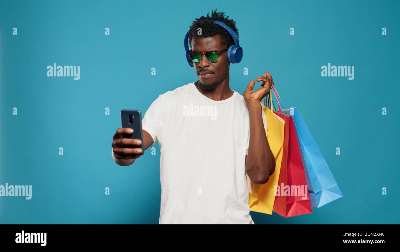 Homme espiègle prenant des photos avec des sacs d'achats sur un smartphone tout en portant des lunettes de soleil et des écouteurs. Adulte moderne prenant des selfies avec le téléphone après l'achat de vêtements dans la vente au détail. Banque D'Images