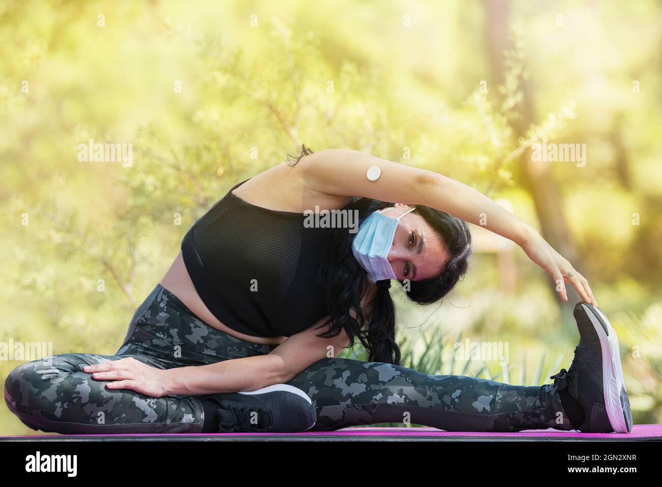 Femme adulte avec vêtements de sport et un capteur de glucose sur son bras qui s'étire sur un tapis violet dans le parc Banque D'Images