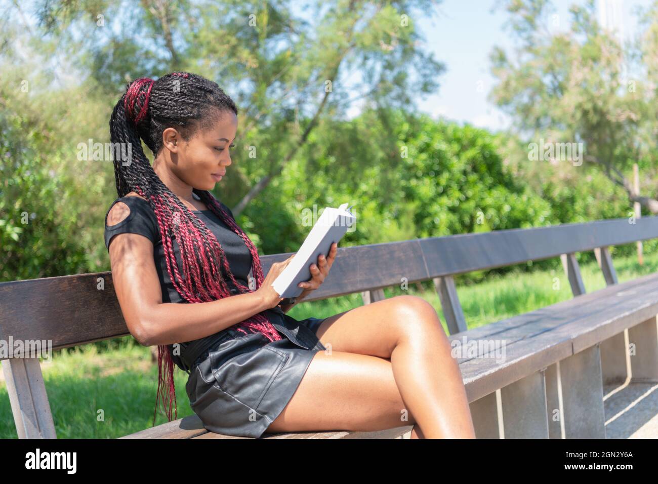 Une femme africaine lisant un livre tout en étant assise dans un banc de parc Banque D'Images