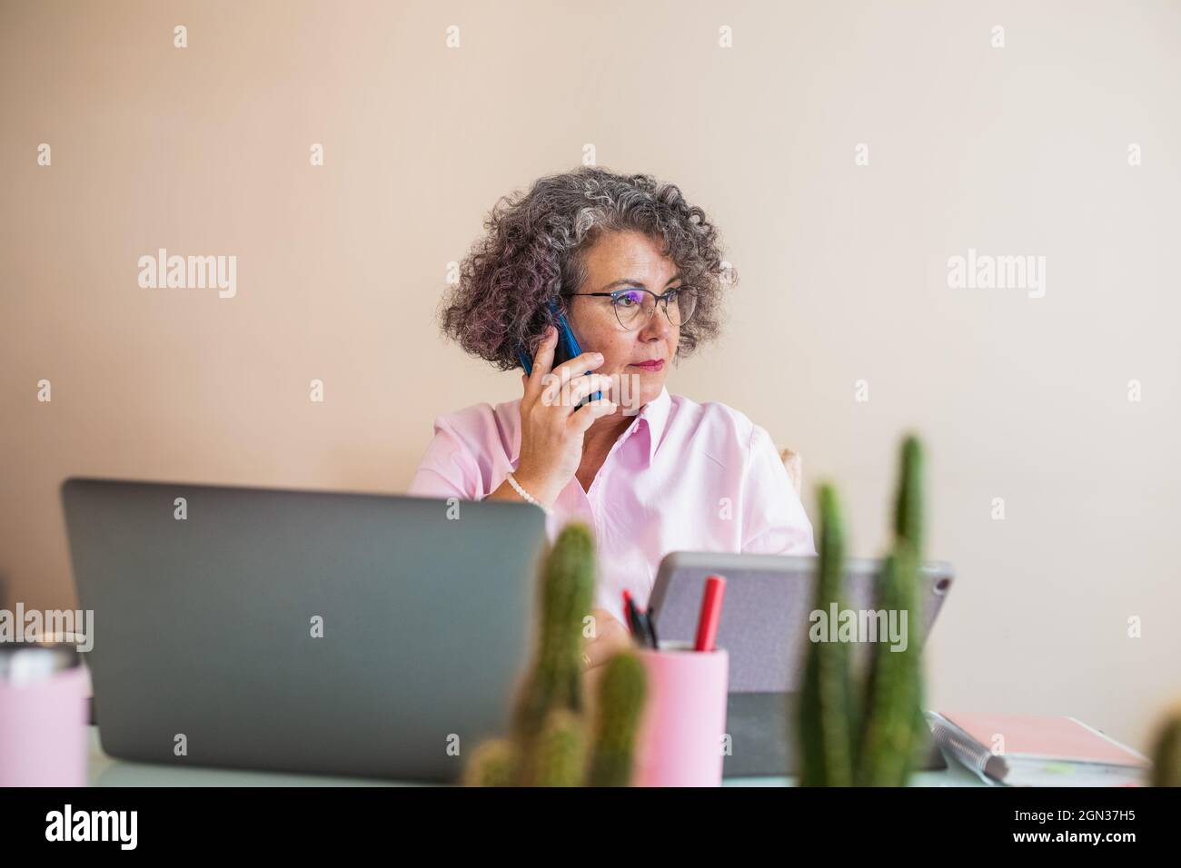 Femme entrepreneure âgée concentrée en lunettes parlant sur un téléphone portable contre un netbook sur une table dans un espace de travail Banque D'Images