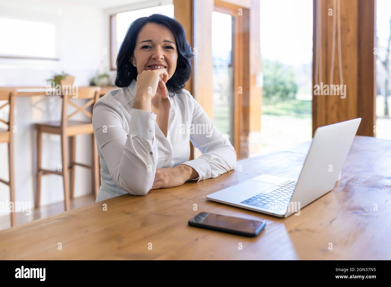 Une femme freelance mûre et heureuse navigue sur Internet sur un netbook en travaillant sur un nouveau projet tout en étant assise à la table à la maison Banque D'Images