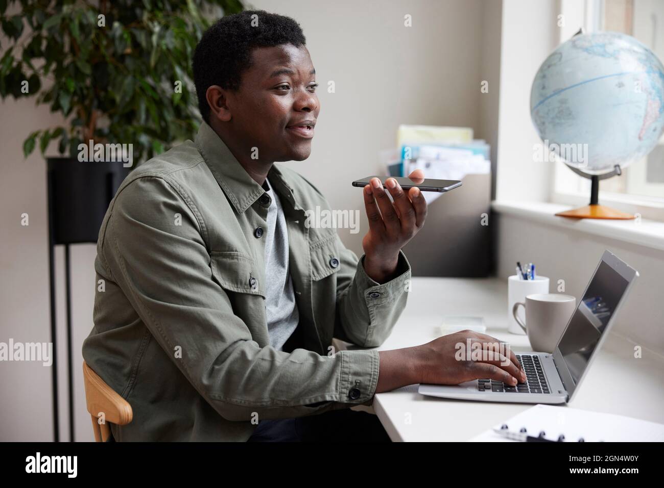 Jeune homme enthousiaste travaillant à domicile sur ordinateur portable parlant dans Mic sur téléphone mobile Banque D'Images