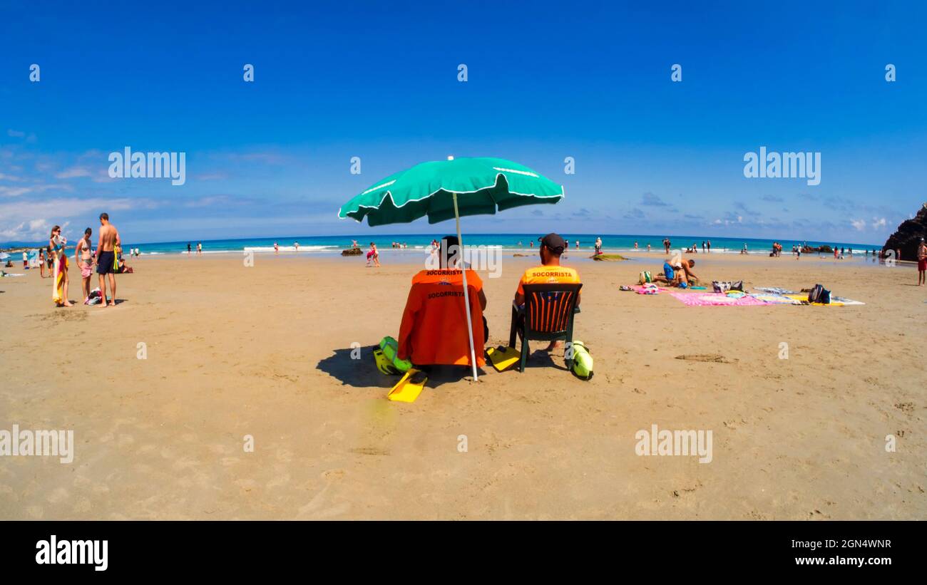 Ribadeo, Galice, Espagne. 07/26/2021 couple de veilleurs de plage assis sous un parapluie sur la plage de Las Catedrales, un ciel bleu par jour d'été. Banque D'Images