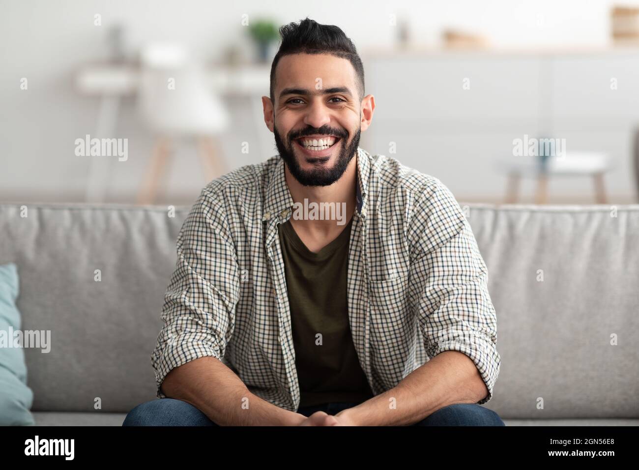 Portrait d'un beau jeune arabe souriant et regardant l'appareil photo, assis sur un canapé à la maison Banque D'Images