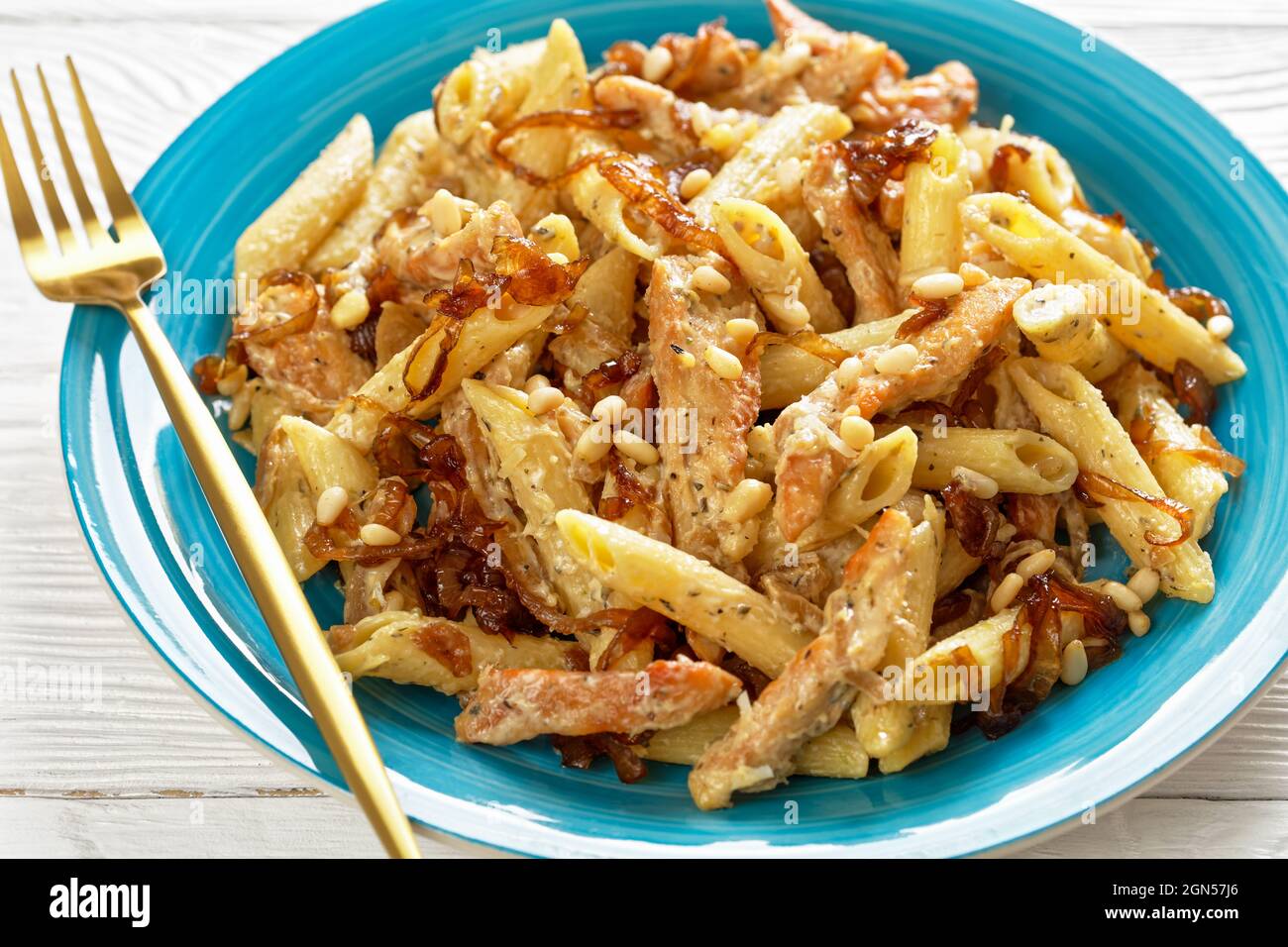 poulet alfredo pâtes penne avec oignon caramélisé arrosé de pignons de pin et parmesan râpé sur une assiette bleue sur une table en bois blanc, gros plan Banque D'Images