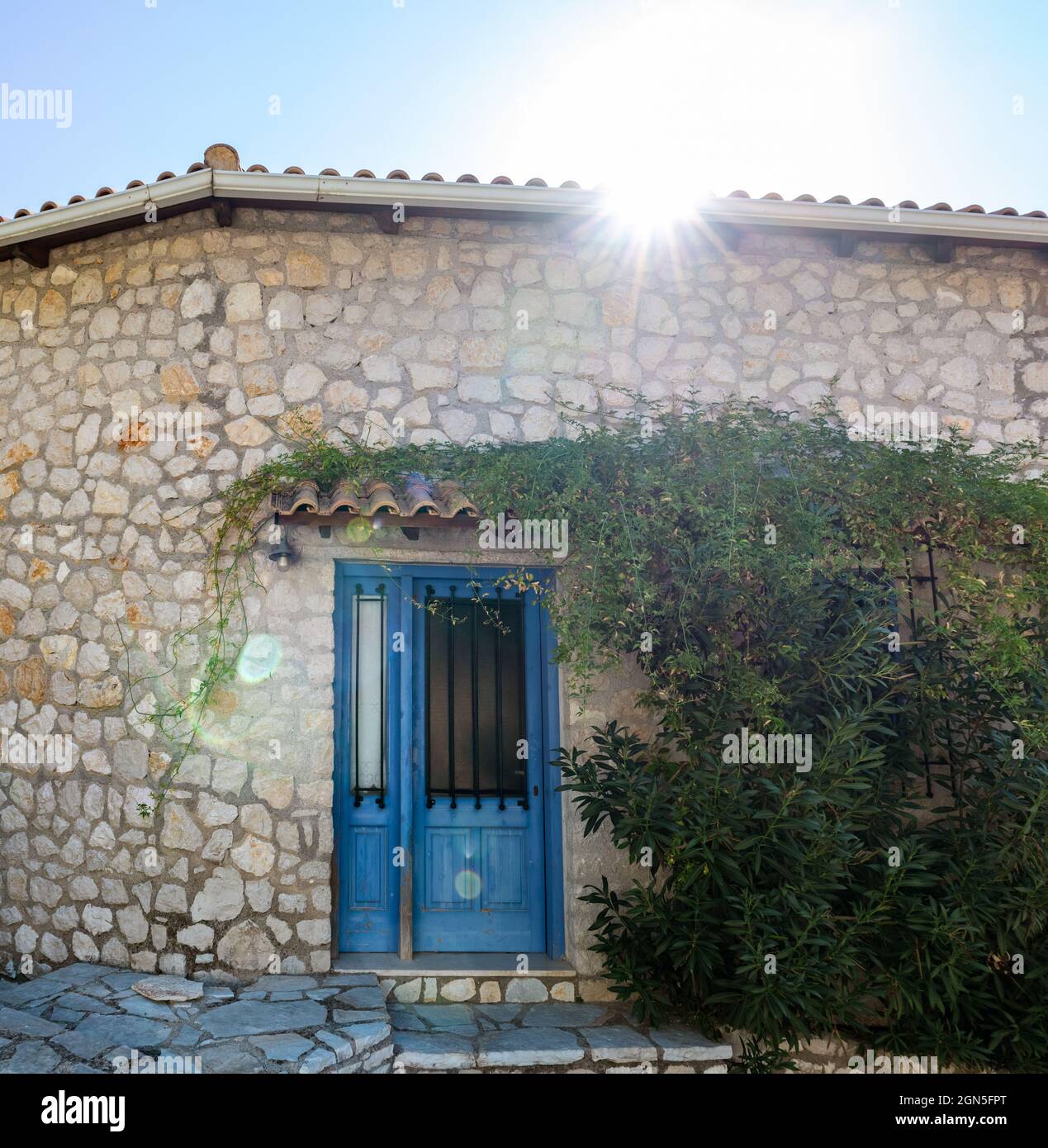 Le soleil brille au-dessus du toit traditionnel grec de la maison avec des murs en pierre, des volets de portes bleues et des branches d'arbres vertes. Voyage d'été en Méditerranée traditionnelle Banque D'Images